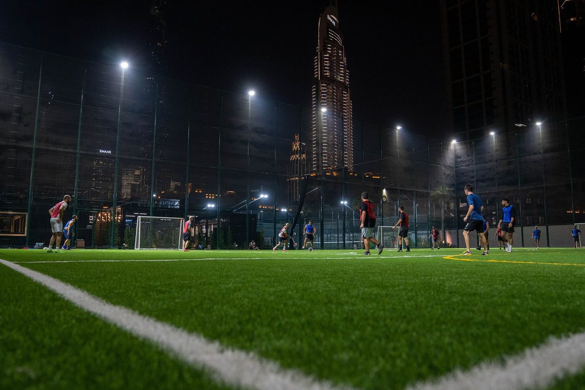 Five-a-side football under floodlights on the Ballers pitch at night, the lit Downtown Dubai towers rising behind the cage