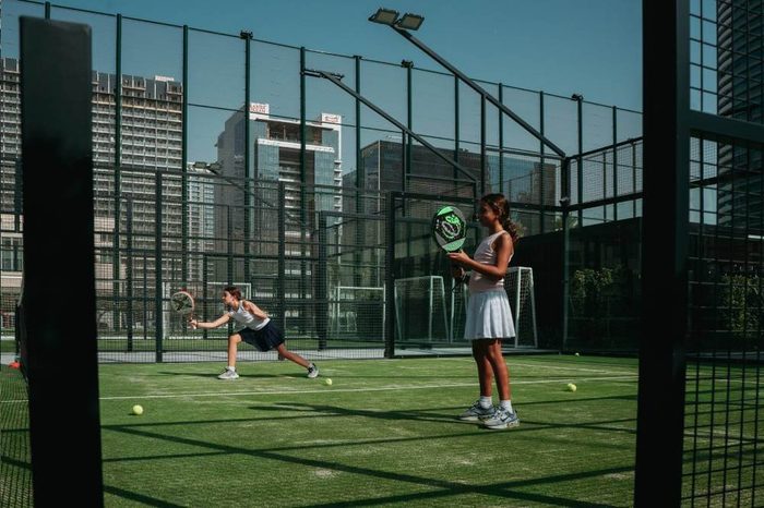 A padel rally in play on a Ballers court, Downtown towers standing behind the glass wall
